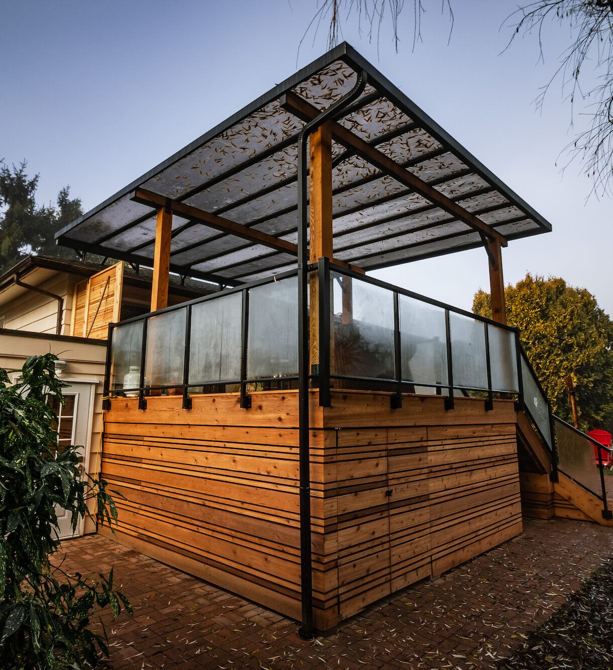Elevated covered cedar deck with horizontal cedar skirting, glass railings, and steel-framed clear roof