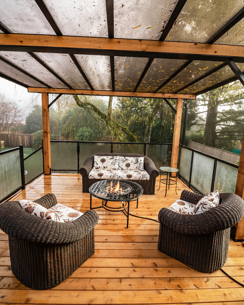Interior view of a covered cedar deck with timber posts, clear roof panels, and a firepit lounge setup