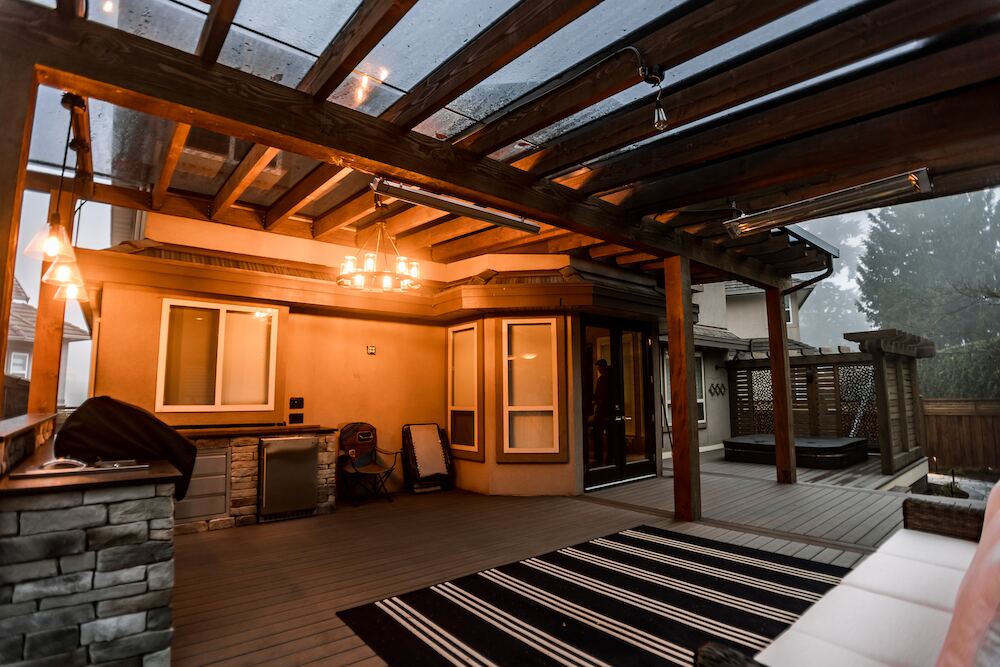 Cedar-framed glass patio cover at dusk with pendant lighting, infrared heaters, and a built-in stone outdoor kitchen