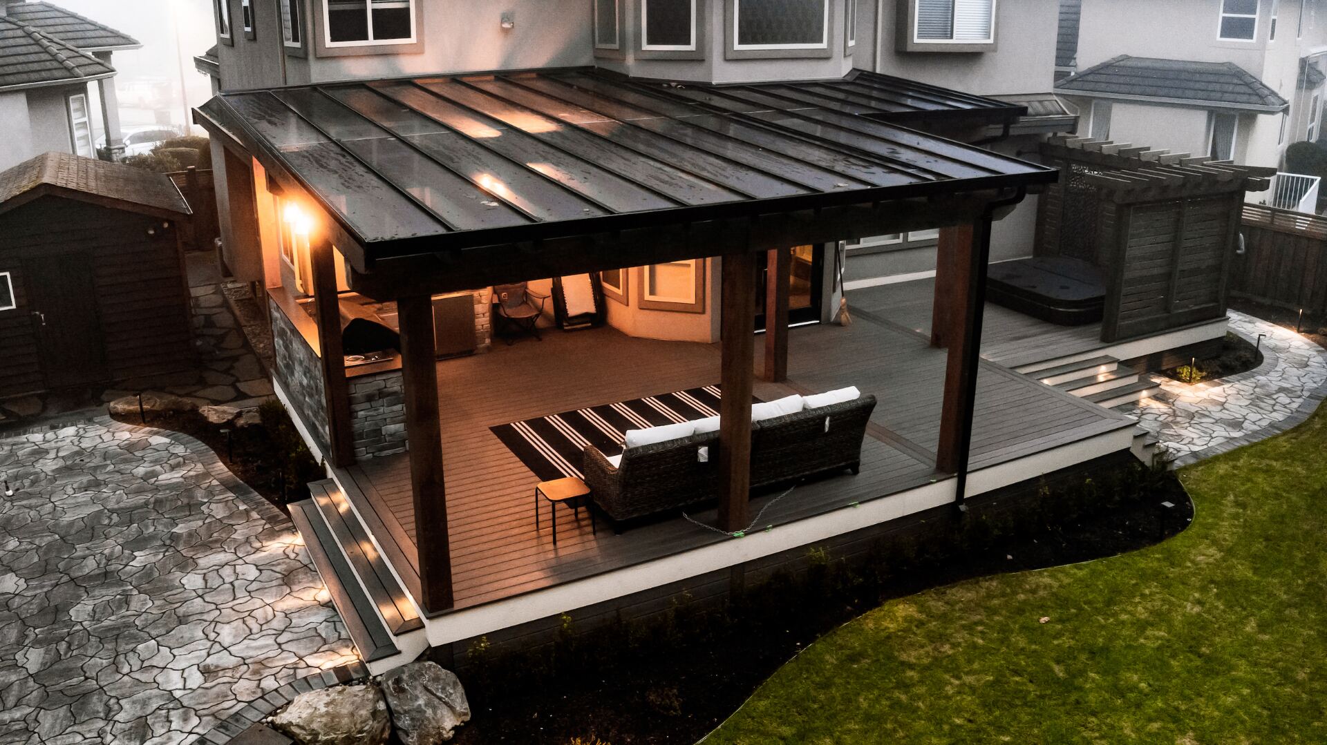 Aerial view of a covered composite deck at dusk with warm interior lighting, glass roof panels, timber posts, and a built-in stone outdoor kitchen