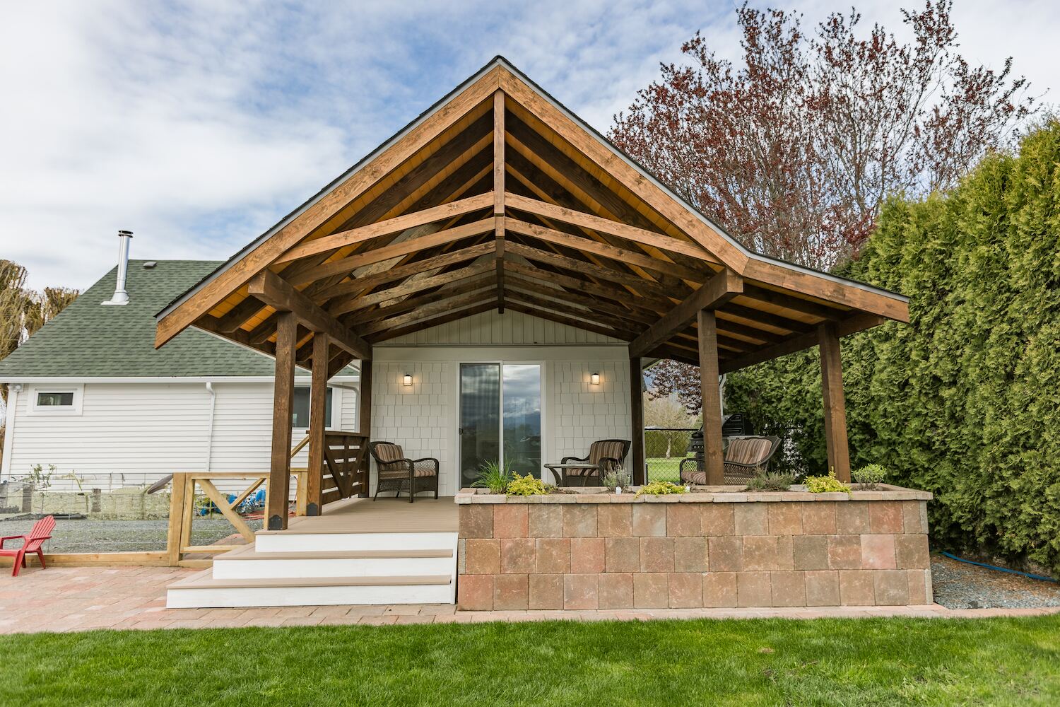 Gabled timber-frame patio cover with exposed cedar trusses, stone planter wall, and covered seating overlooking a Fraser Valley backyard