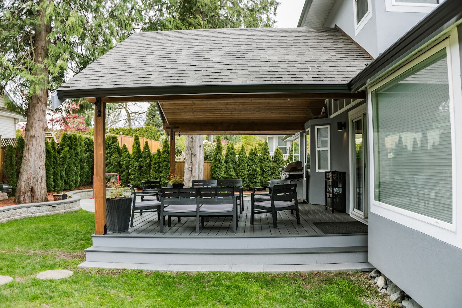 Hip-roof patio cover with shingle roofline matching the home, covering a grey deck and outdoor dining set